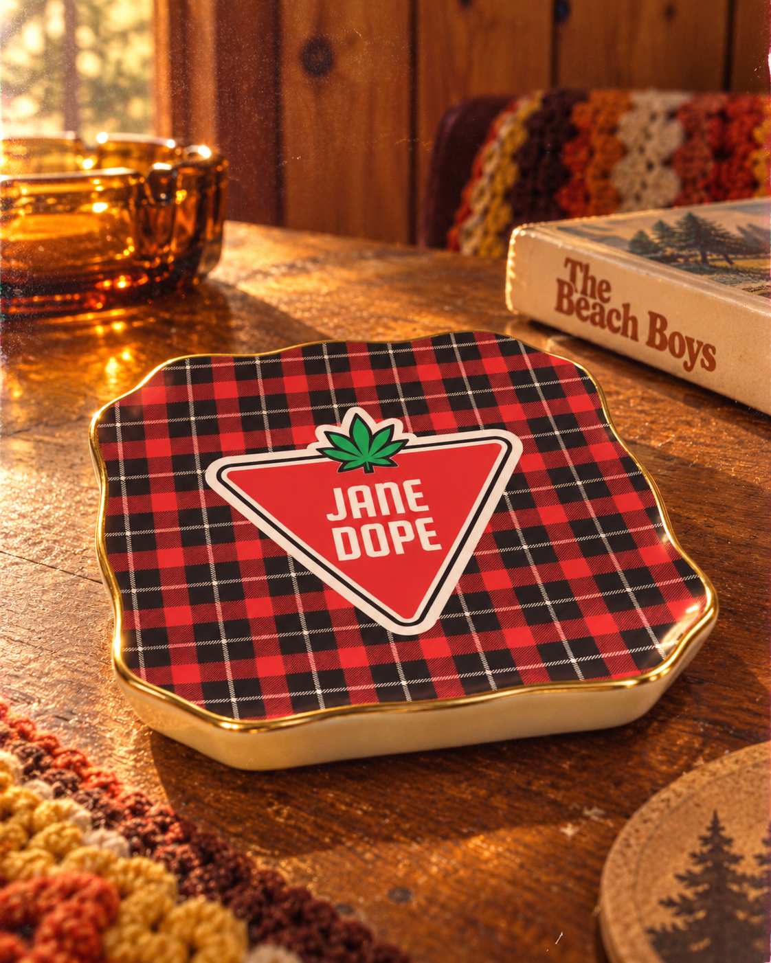 A ceramic coasters featuring a red and black buffalo plaid pattern with a central red triangle logo that reads 'JANE DOPE' with a green leaf above it, set on a rustic wooden table.