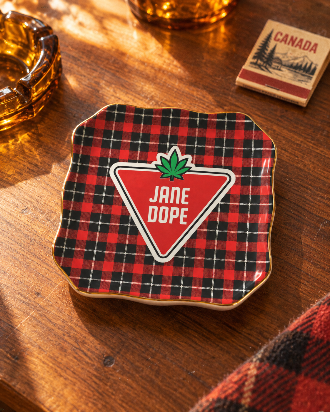 A square enamel pin with a red and black plaid pattern, featuring a central red triangle with a green cannabis leaf and the text 'JANE DOPE' in white. The pin is displayed on a wooden table next to a matchbook and amber glass.