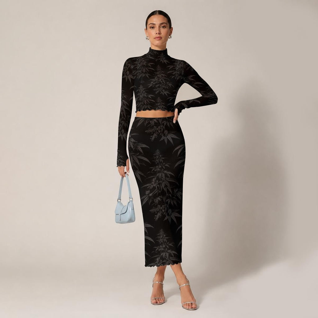 A model stands against a plain beige studio background wearing a matching black long-sleeve crop top and high-waisted midi skirt set with a dark gray botanical pattern. She is holding a small light blue handbag and wearing clear high-heeled sandals.