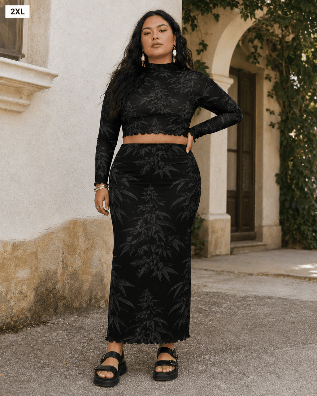 A woman modeling a black two-piece set consisting of a long-sleeve crop top and a high-waisted maxi skirt with a tonal botanical pattern, set against an outdoor stone wall background.