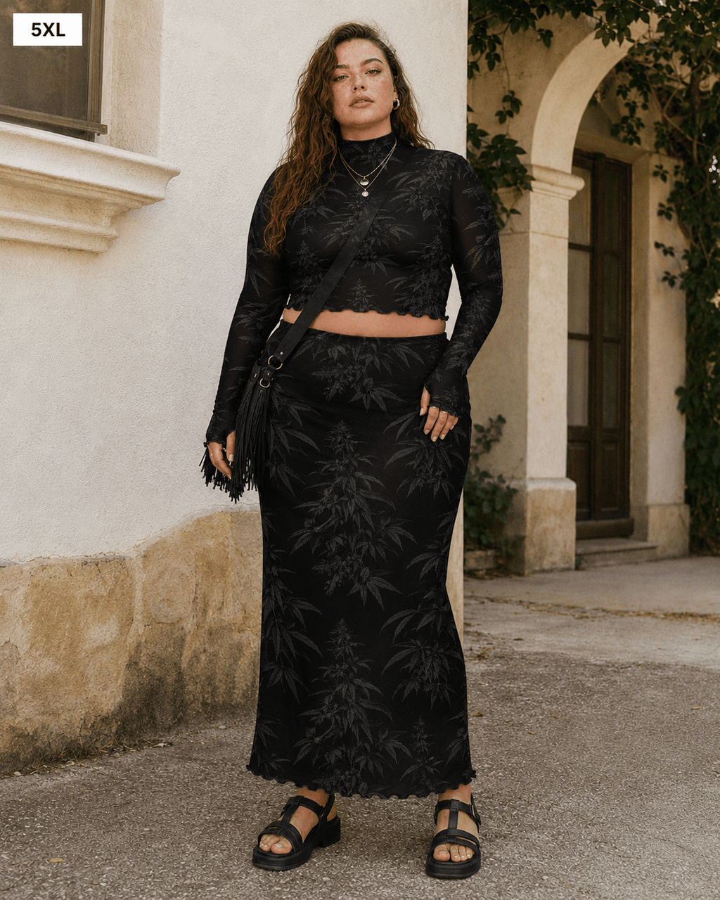 A woman modeling a black two-piece lace set consisting of a long-sleeve crop top and a matching maxi skirt, standing outdoors in front of a beige stucco building.