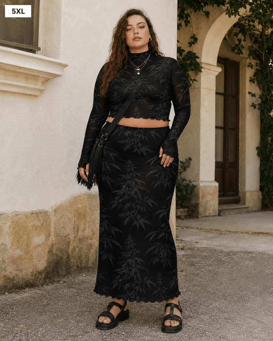 A woman modeling a black two-piece lace set consisting of a long-sleeve crop top and a matching maxi skirt, standing outdoors in front of a beige stucco building.