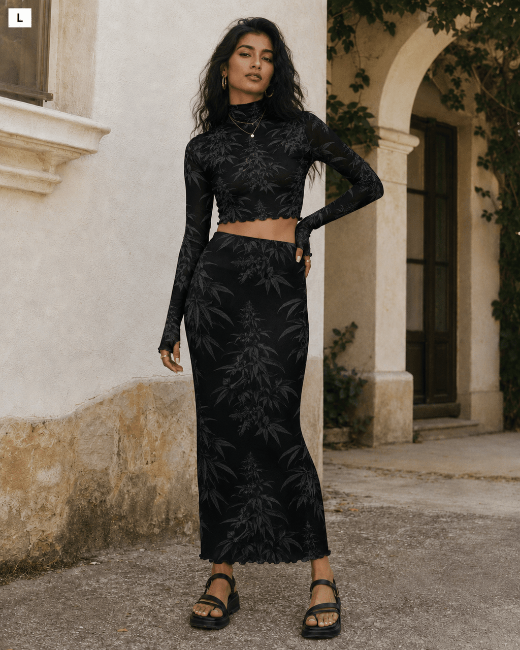 A woman modeling a black two-piece set featuring a long-sleeve crop top and a matching high-waisted maxi skirt with a tonal botanical leaf pattern, set against an outdoor stone wall and archway background.