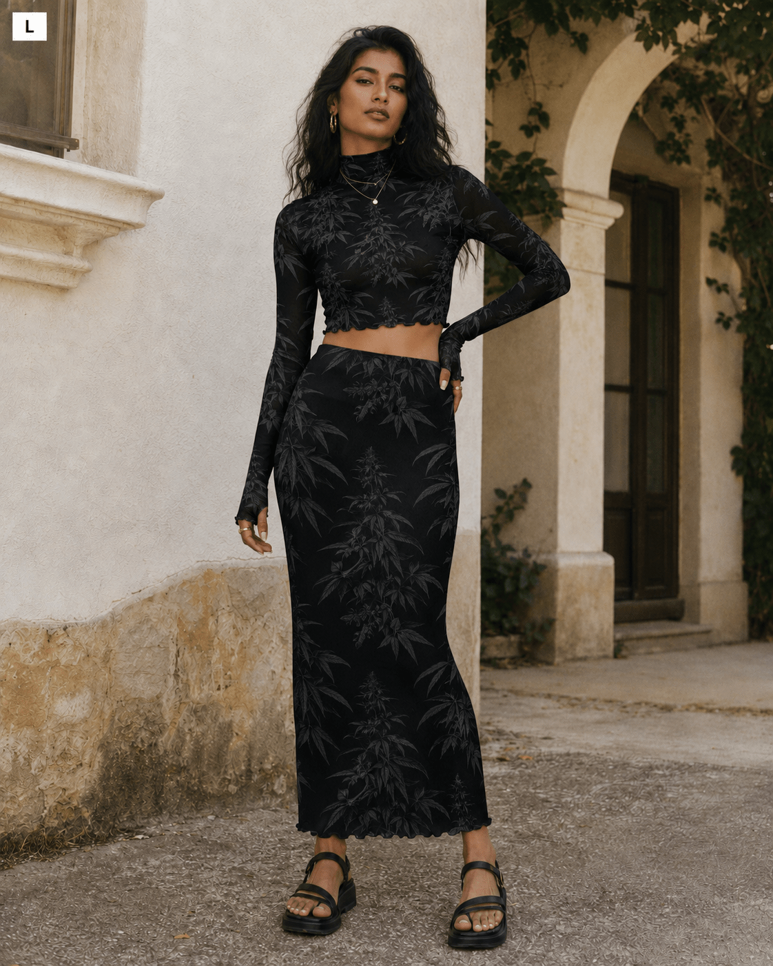 A woman modeling a black two-piece set featuring a long-sleeve crop top and a matching high-waisted maxi skirt with a tonal botanical leaf pattern, set against an outdoor stone wall and archway background.