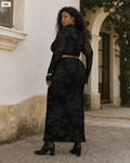 A woman modeling a black two-piece lace set consisting of a long-sleeve crop top and a matching maxi skirt, standing outdoors in front of a beige stucco building.