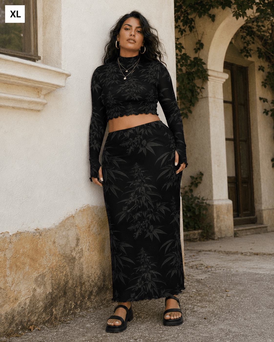 A woman modeling a black two-piece lace set consisting of a long-sleeve crop top and a matching maxi skirt, standing outdoors against a beige stucco wall.