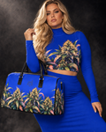 A woman models a royal blue matching two-piece outfit with a high-neck long-sleeve crop top and high-waisted skirt, both featuring a large cannabis plant print. She is holding a matching blue duffel bag with black handles and trim, set against a dark studio background.