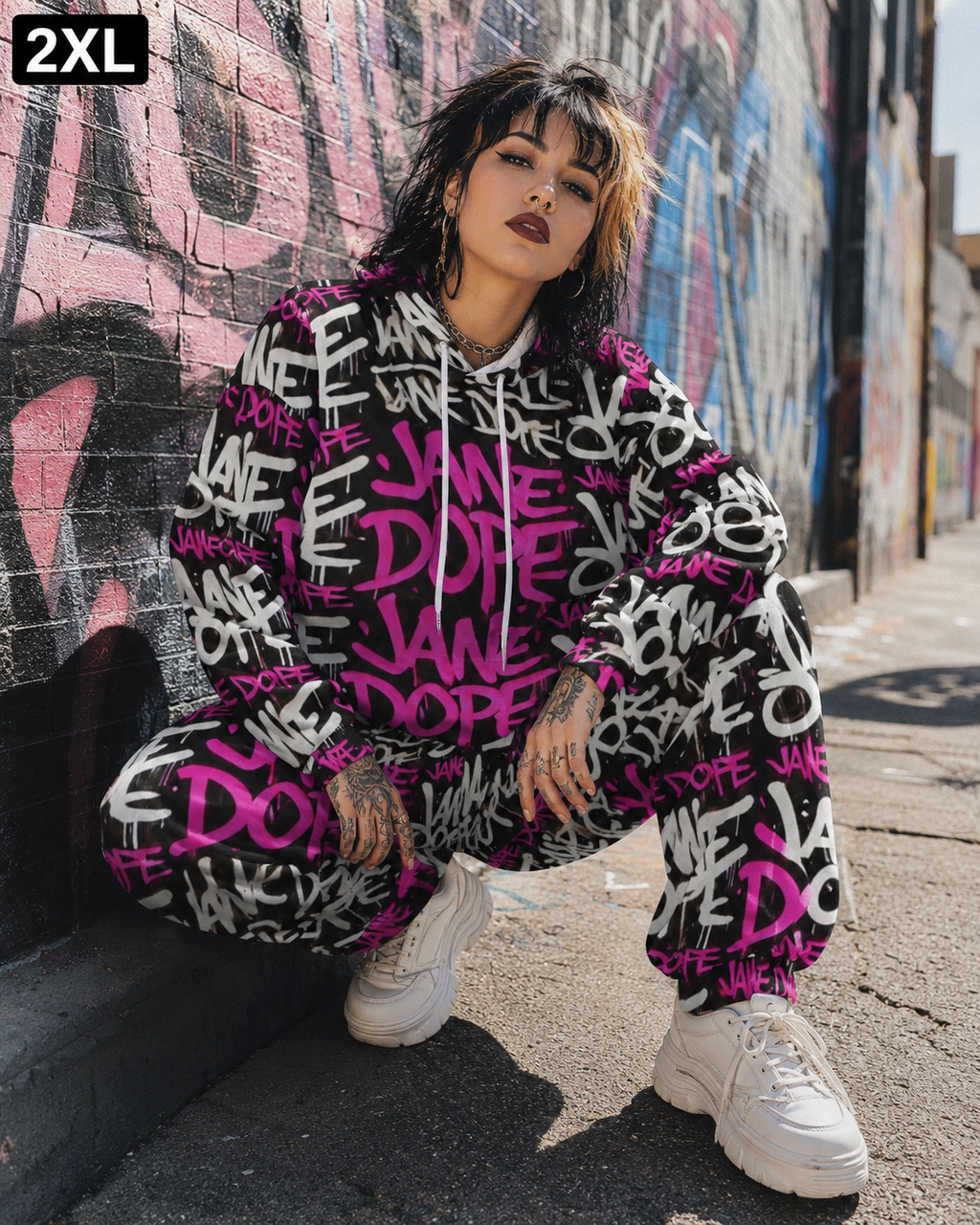 A model crouching outdoors against a graffiti-covered wall, wearing a matching oversized hoodie and sweatpants set covered in a bold black, white, and hot pink graffiti text print. The image has a '2XL' size label in the top-left corner.