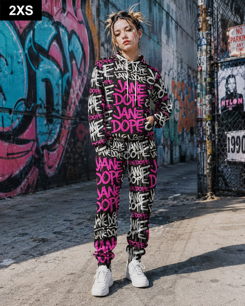 A woman stands on a city sidewalk in front of a graffiti-covered wall, wearing a matching black hoodie and jogger pants set covered in bold pink and white 'JANE DOE' graffiti-style text. She is also wearing white sneakers.