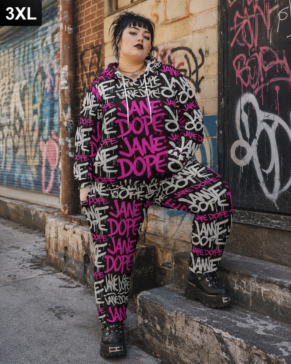A person wearing a matching oversized hoodie and jogger pants set with a bold graffiti-style print featuring the words 'JANE DOPÉ' in bright pink, black, and white, sitting on concrete steps against a graffiti-covered wall.