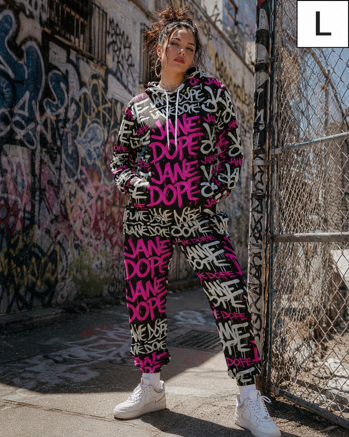 A model wearing a matching black and pink graffiti sweatshirt and jogger pants set, standing in an urban alleyway with graffiti walls and a chain-link fence. A size 'L' label is visible in the top right corner.