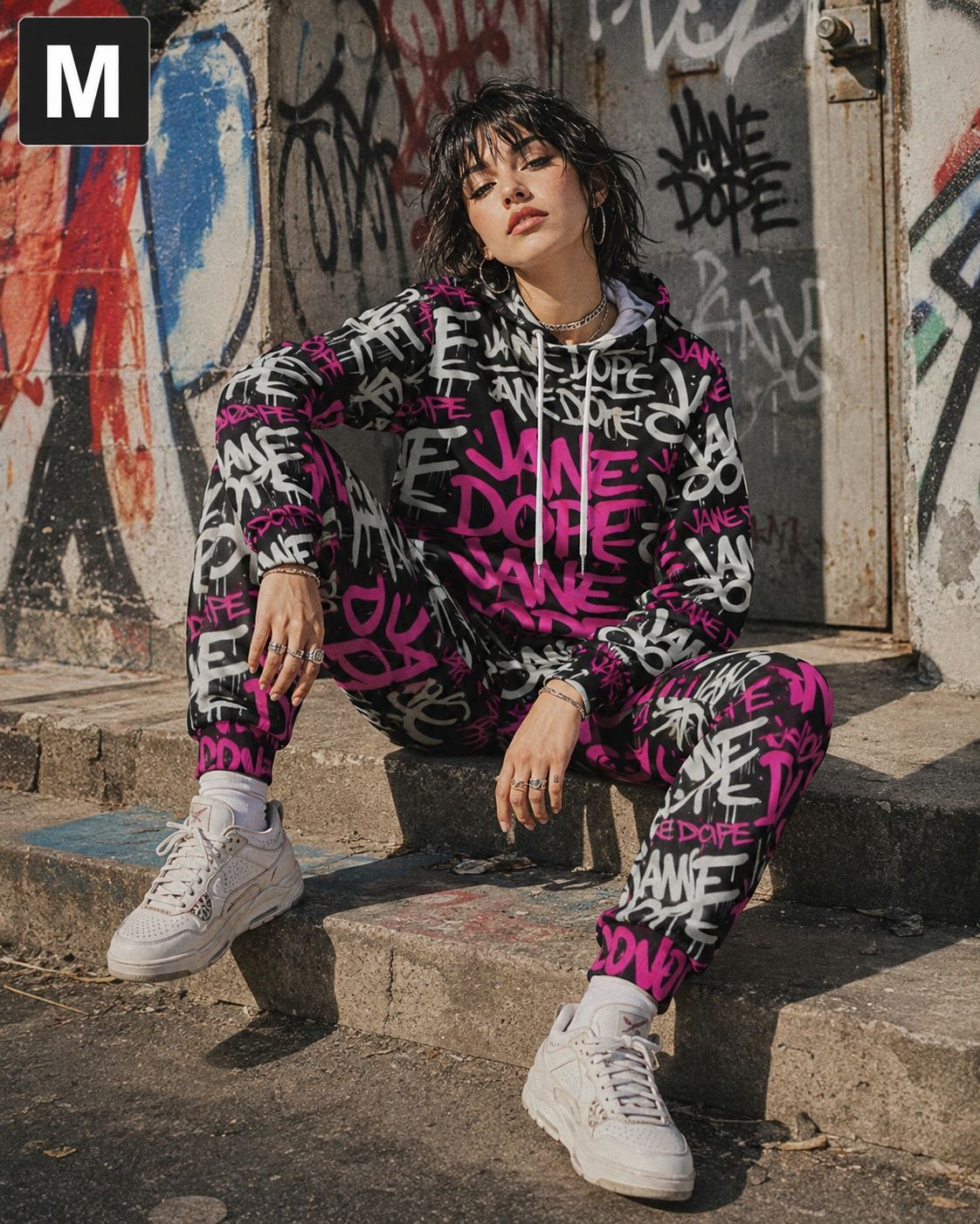 A model wearing a matching pink, black, and white graffiti print hoodie and sweatpants set, sitting on concrete steps in front of a graffiti-covered wall.