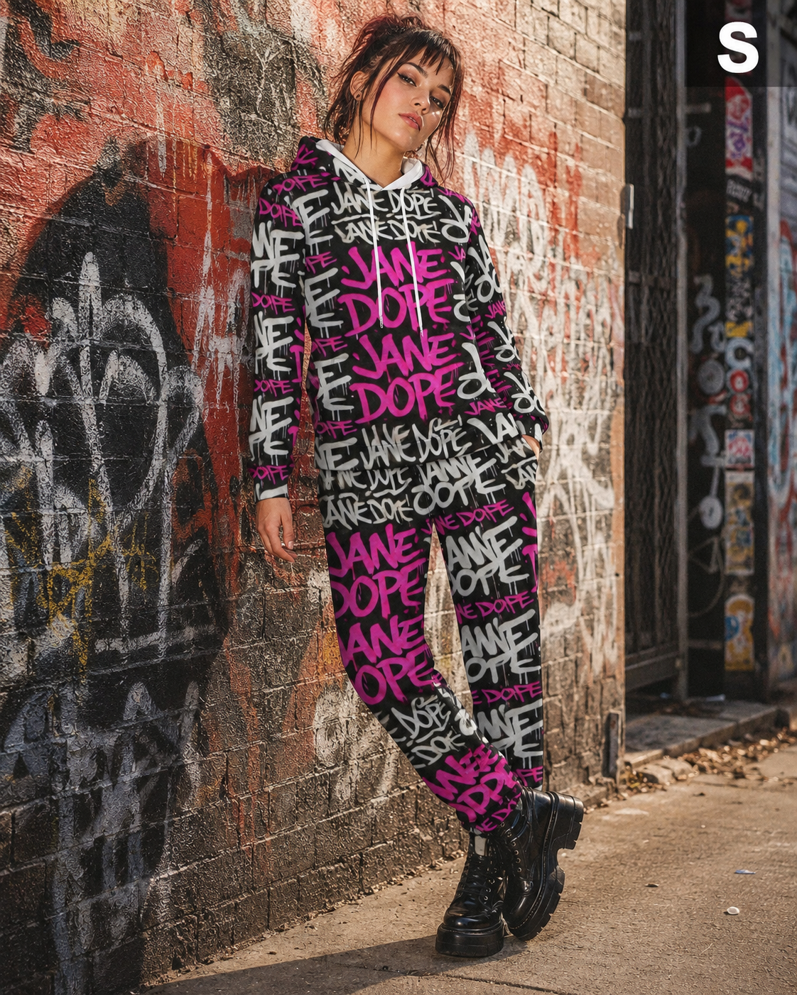 A woman stands against a graffiti-covered brick wall wearing a matching black and pink graphic sweatshirt and jogger pants set with white text, paired with black combat boots.
