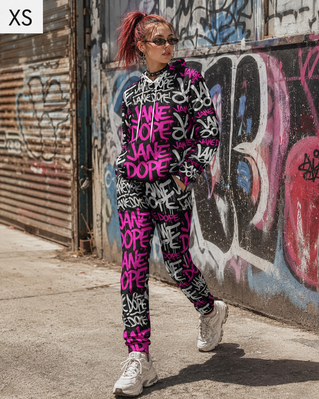 A person wearing a matching black sweatshirt and jogger pants set with a bright pink and white graffiti print, standing outdoors against a graffiti-covered wall. The sweatshirt features the text 'JANE DOPE' repeatedly in pink and white, and the joggers have a similar pattern.