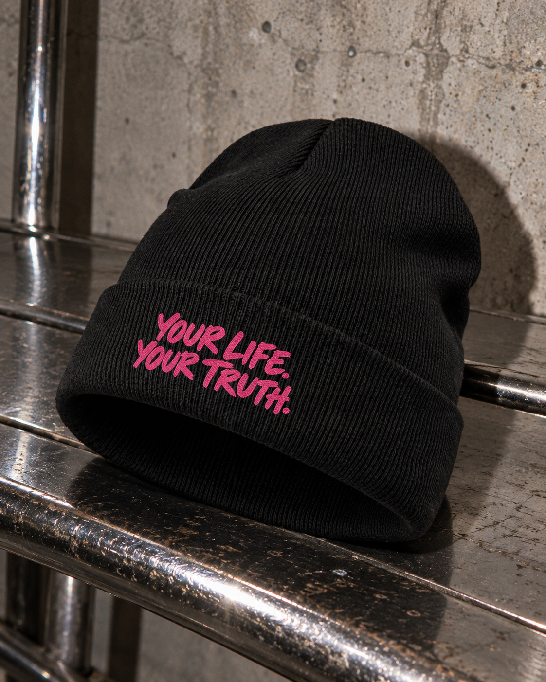 Black BE JANE DOPE knit beanie with pink YOUR LIFE. YOUR TRUTH. stitching resting on a chrome bench.
