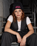 Black BE JANE DOPE stitch beanie with pink slogan, worn with a tailored vest in a seated stairwell flash editorial.