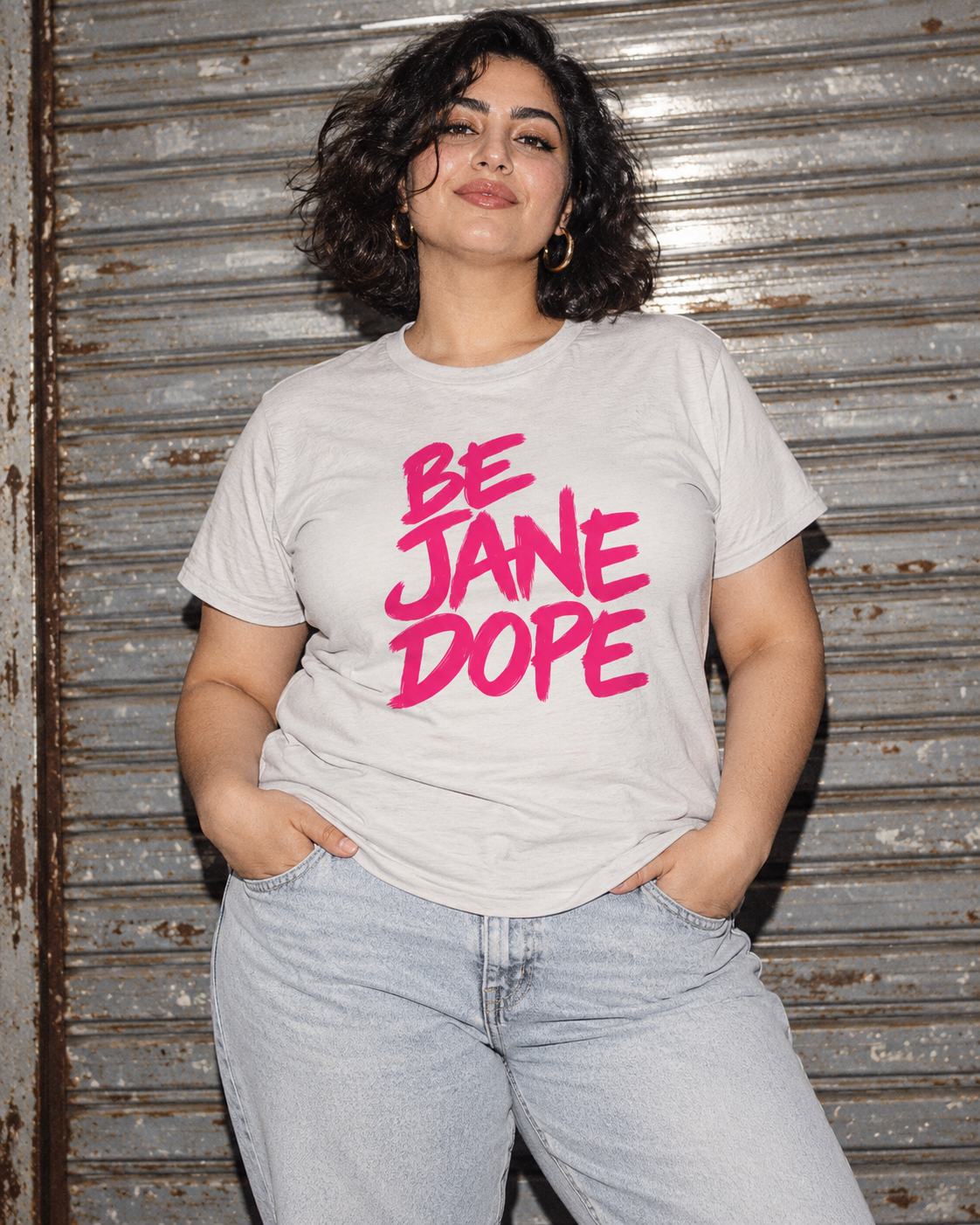Front of BE JANE DOPE cotton tee with pale jeans against a metal shutter.