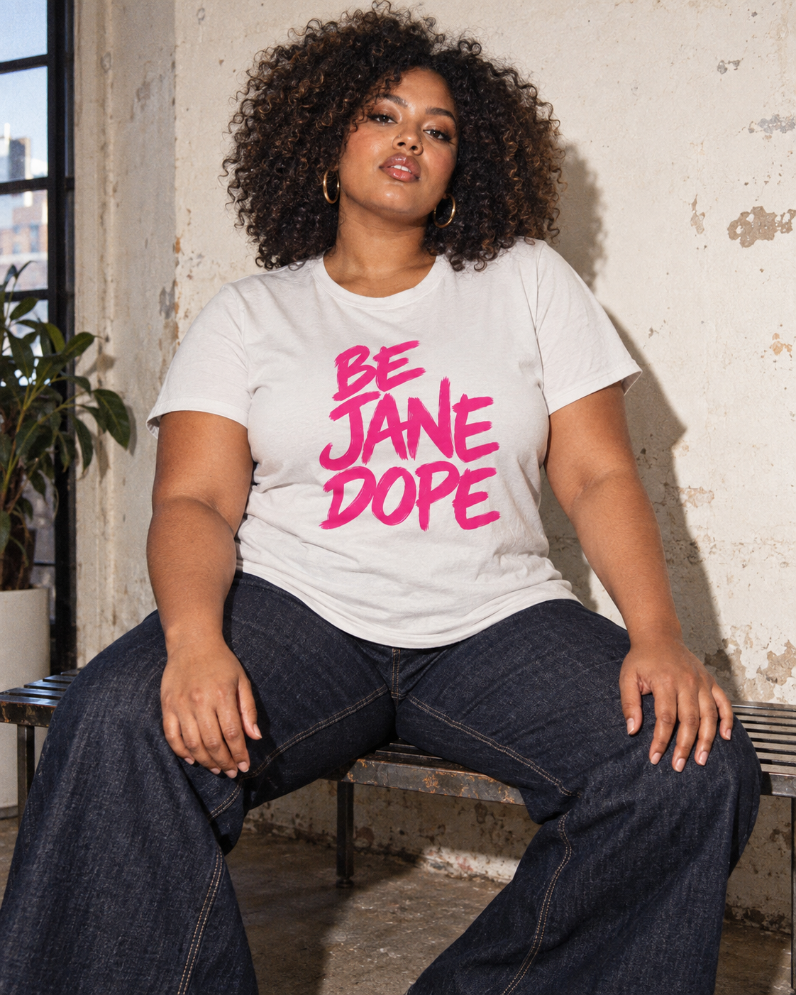 BE JANE DOPE tee styled with dark flared jeans, seated on a metal bench.