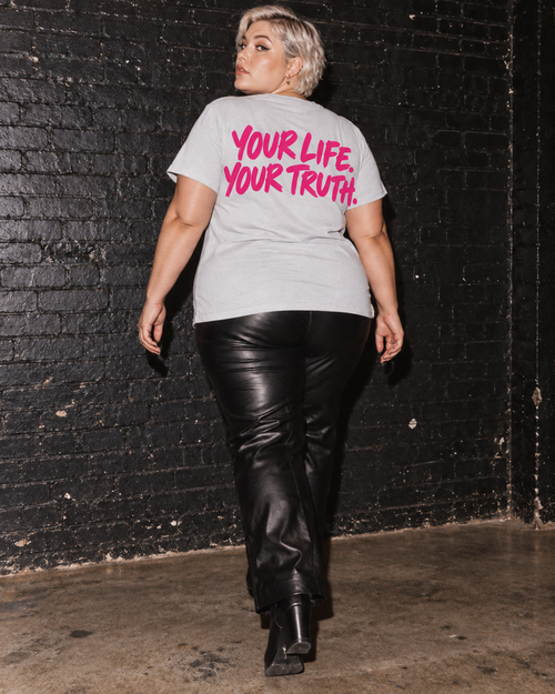 Back of BE JANE DOPE tee with black trousers against a dark brick wall.