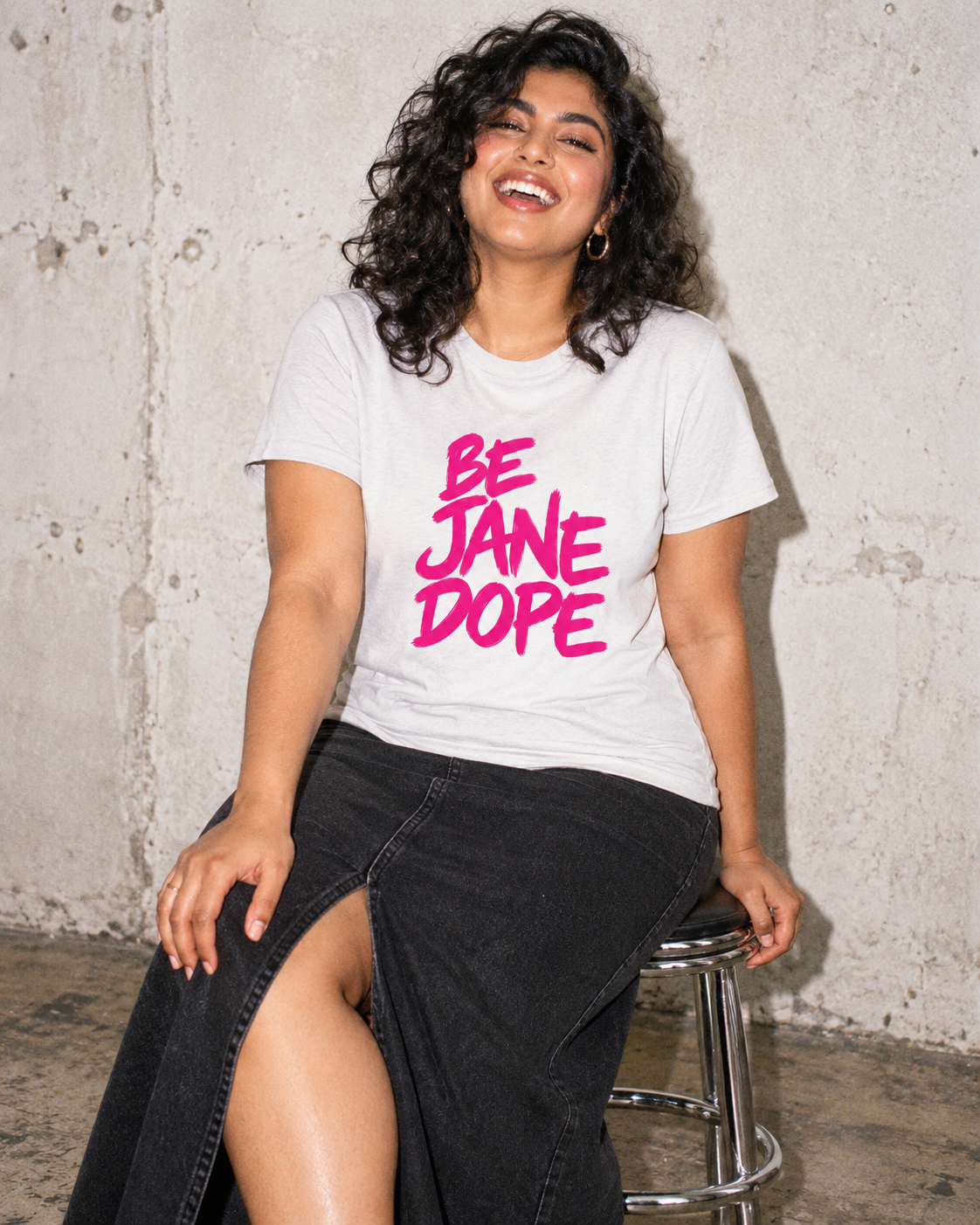 BE JANE DOPE graphic tee styled with a black denim skirt, seated on a chrome stool.