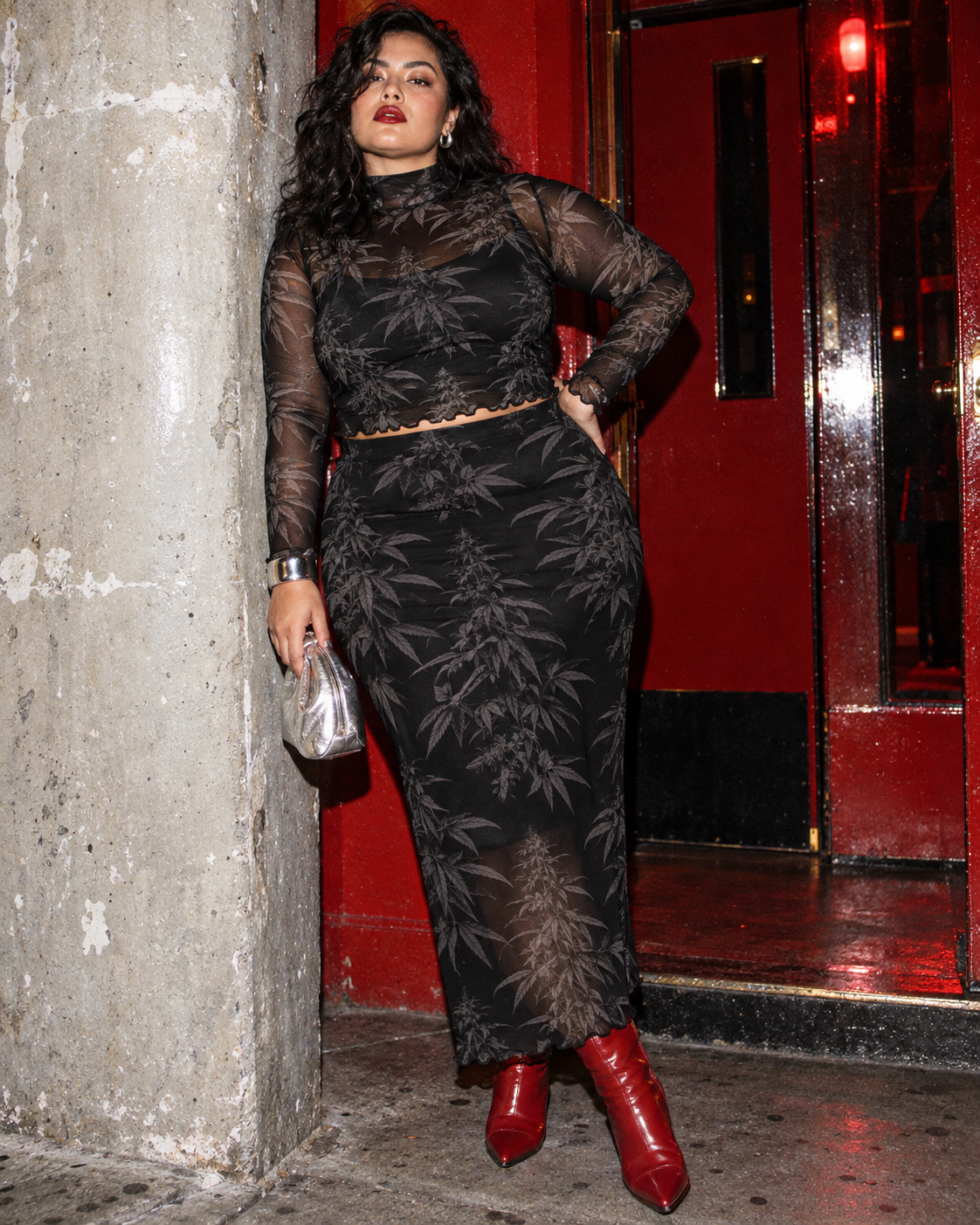 Black Widow botanical mesh set styled with red boots and a silver bag beside a concrete pillar.