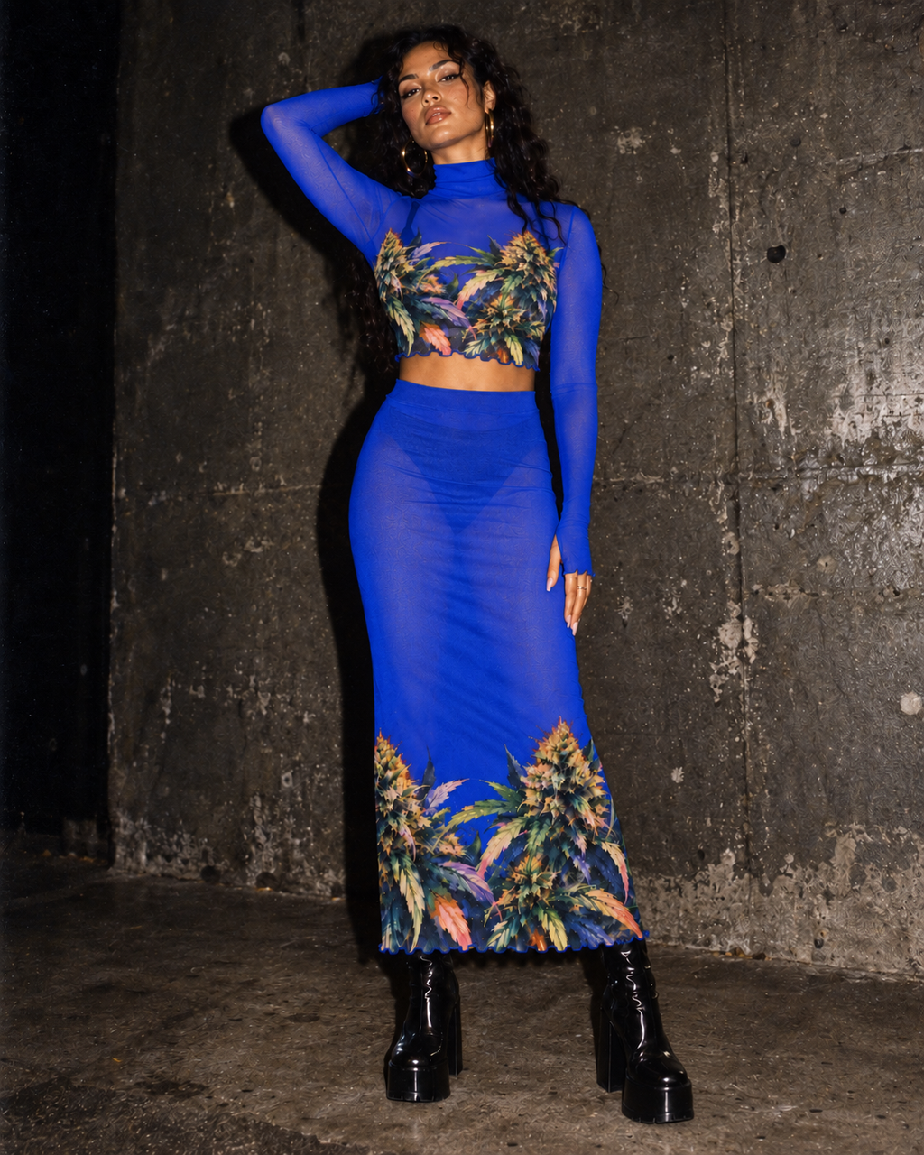 Blue Mazar sheer top and skirt, front view with one arm raised against a dark concrete wall.