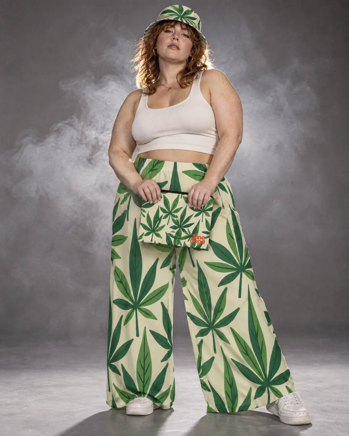 Model in Grass wide-leg pants and bucket hat holding the matching pouch in front, with softly lit smoke behind her.