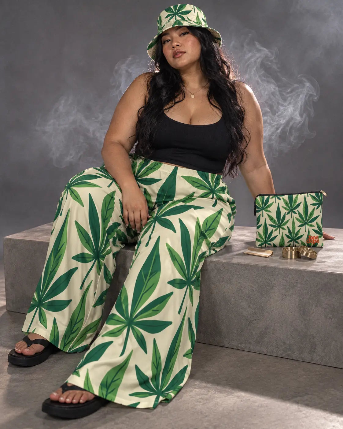 Seated model in the Grass Kit beside its matching pouch, rolling papers and a grinder on a gray studio bench.