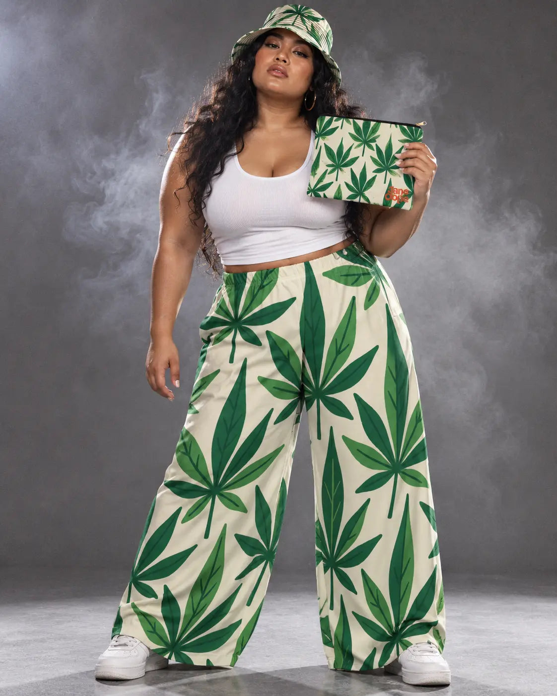 Model in Grass leaf-print pants and bucket hat holding the matching Jane Dope pouch at shoulder height.
