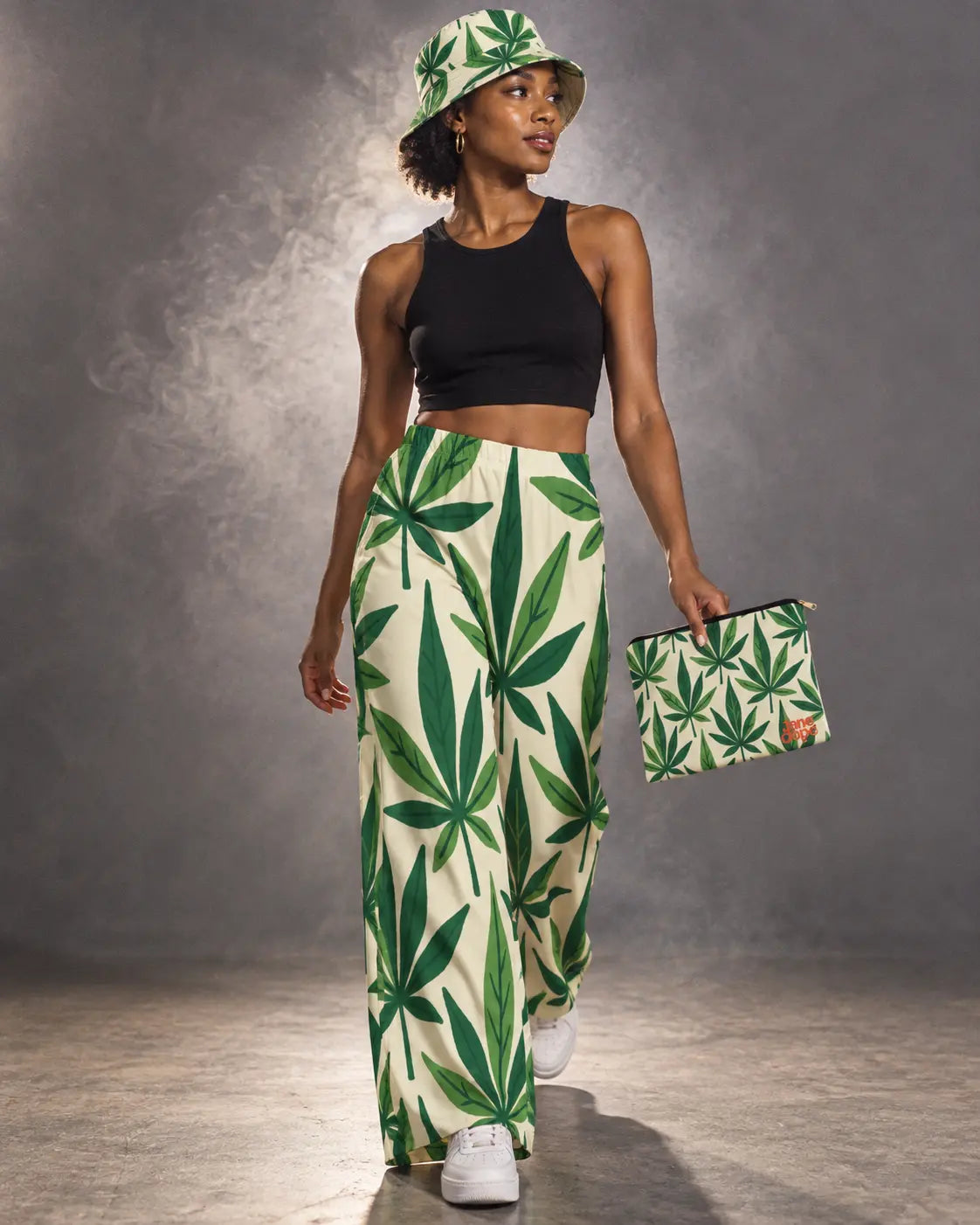 Model walking in the Grass Kit, wearing leaf-print pants and bucket hat while carrying the matching zip pouch.