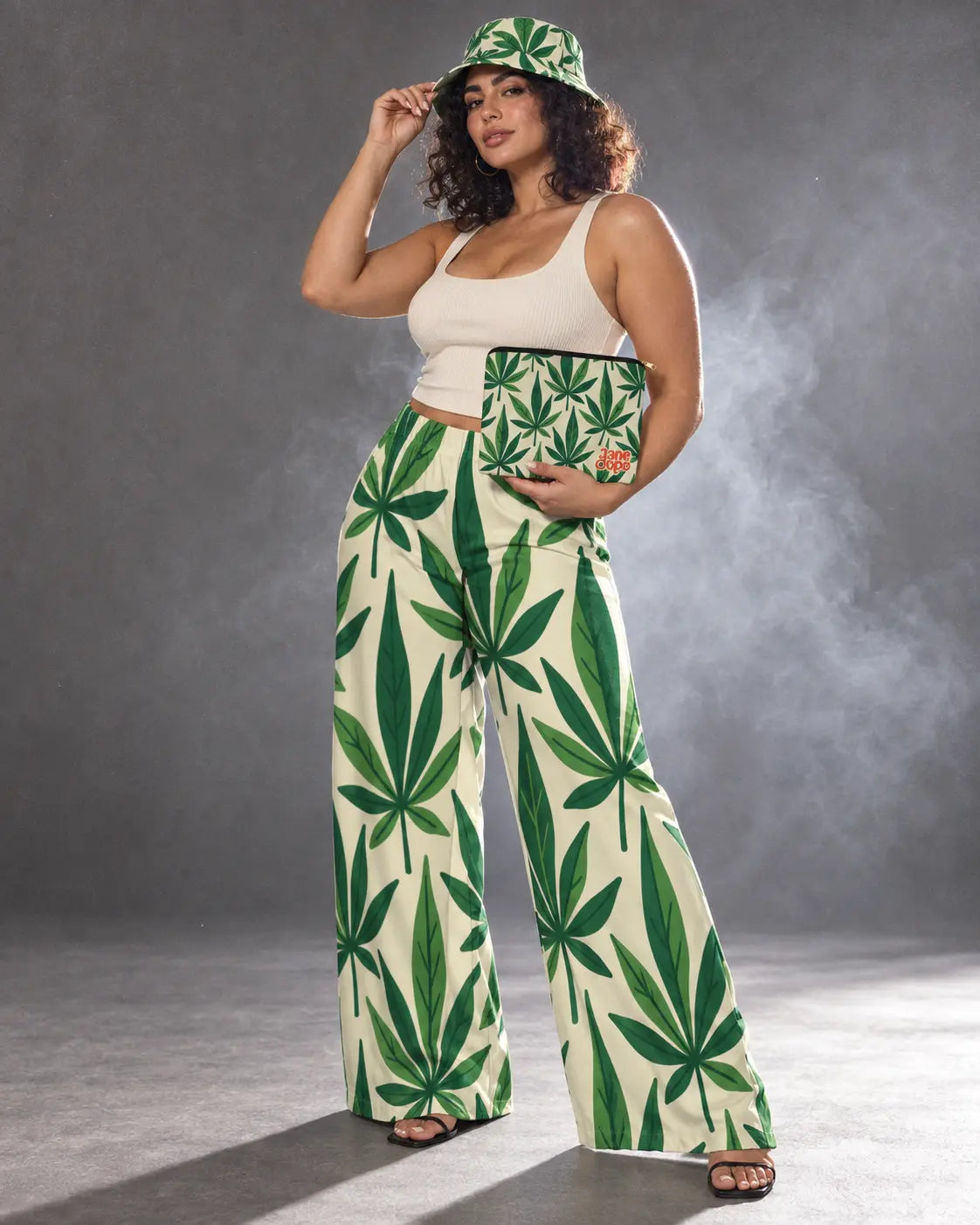 Model touching the Grass bucket hat brim while holding the matching stash pouch at her hip above leaf-print pants.
