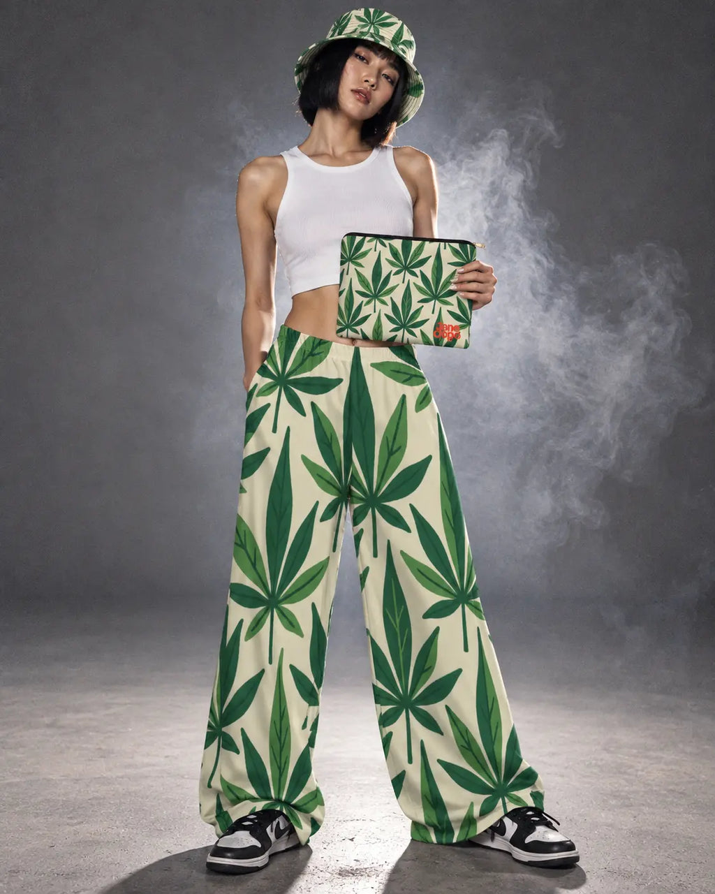 Model standing in Grass wide-leg pants and bucket hat, holding the matching Jane Dope stash pouch against a smoky gray studio backdrop.