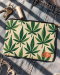 Jane Dope Grass zip pouch with green HER.B leaves and orange logo on faded denim beside keys and lip balm.