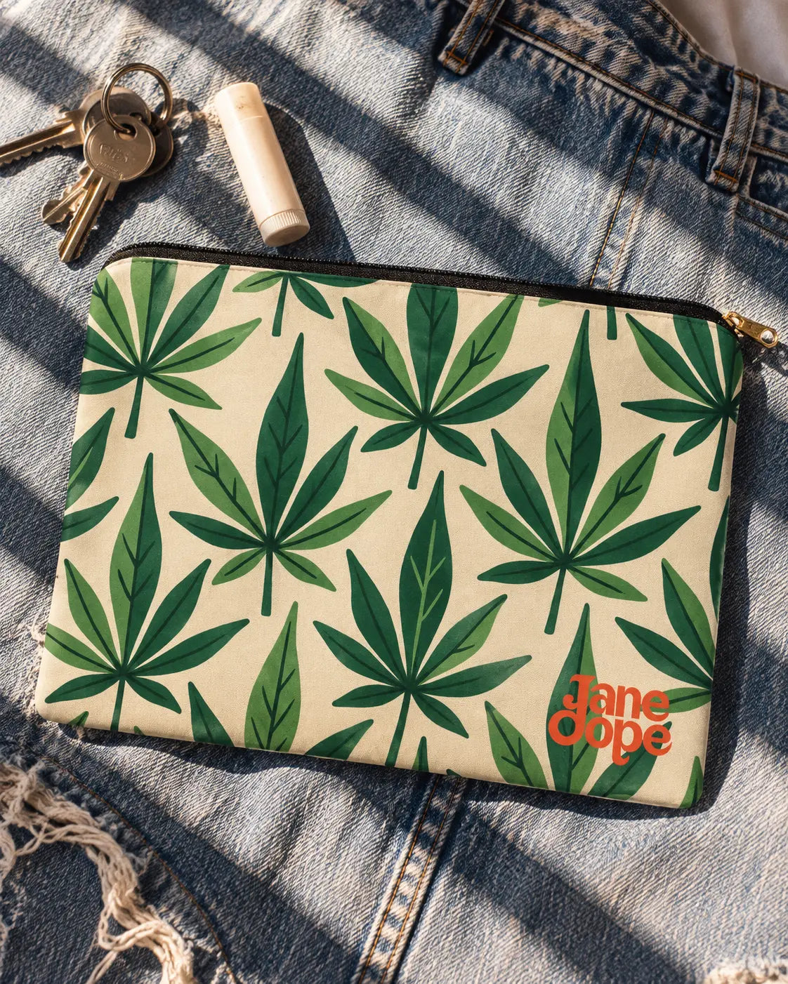 Jane Dope Grass zip pouch with green HER.B leaves and orange logo on faded denim beside keys and lip balm.