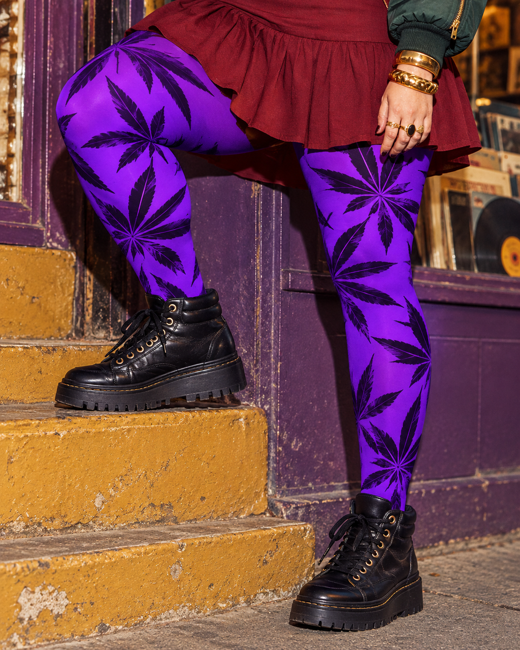 Side detail of Purple Runtz tights with one knee bent on a step, burgundy skirt, gold jewellery and black boots.