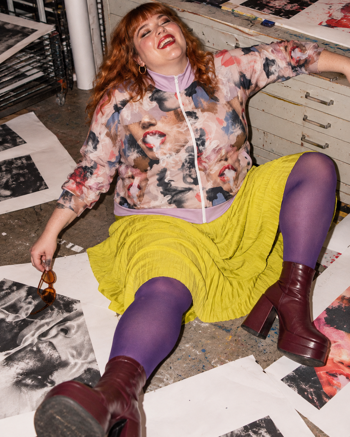 Squad jacket editorial mockup, model laughing among prints on the floor in a yellow skirt and purple tights.