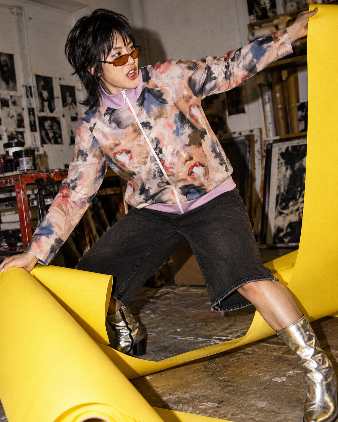 Squad jacket editorial mockup, model pulling yellow paper across a print studio in silver boots.
