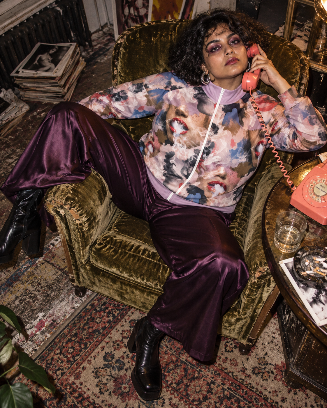 Squad jacket editorial mockup, model reclining in a velvet chair with plum trousers and a coral telephone.